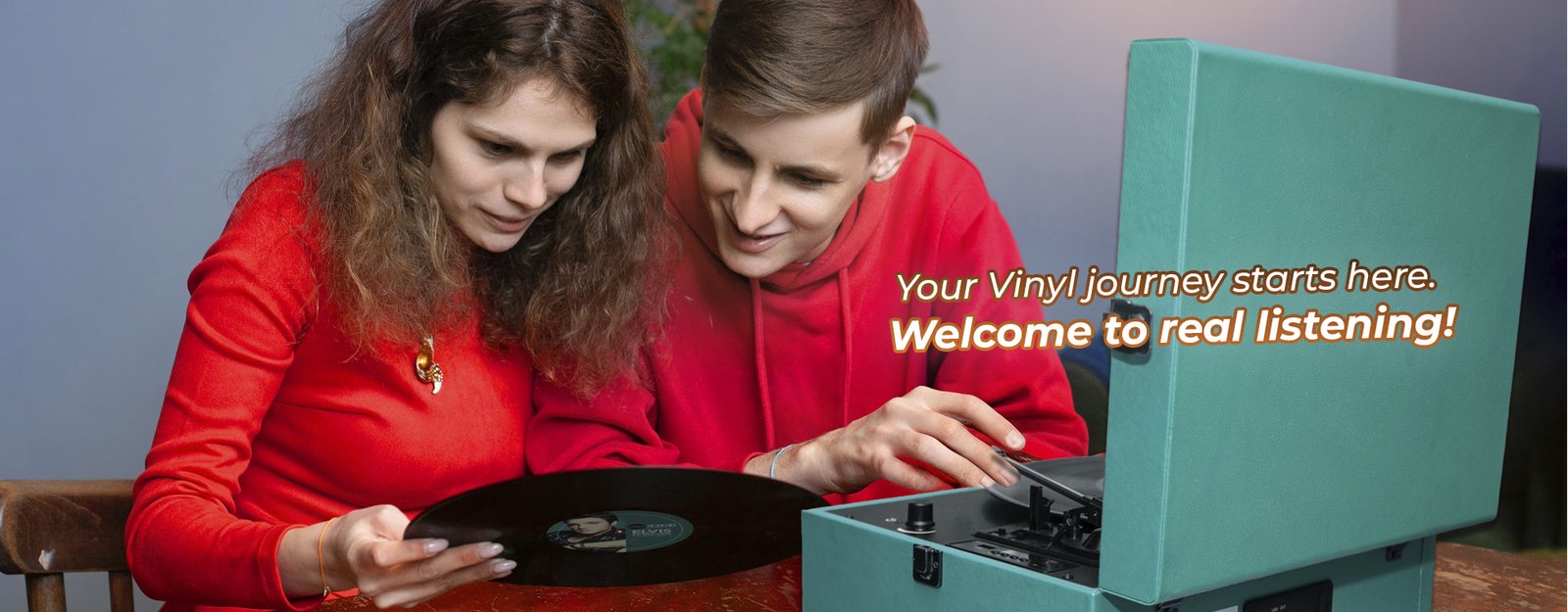 Young couple starting their vinyl journey with a Vinyl Buddy turquoise record player, smiling while placing a record onto the turntable, with the tagline 'Your Vinyl journey starts here. Welcome to real listening!'